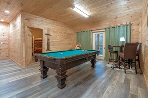 A wooden interior with a pool table, chairs, a table with a lamp, and a door leading to a storage area. - Hot Tub & Dolly Tickets - Gatlinburg Cabin Retreat (Gatlinburg)