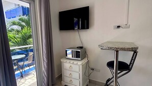 Ein Zimmer mit an der Wand befestigtem Fernseher, einem kleinen Tisch mit Stuhl, einer weißen Kommode und einer Schiebetür zu einem Außenbereich mit Stühlen und einem blauen Zaun.