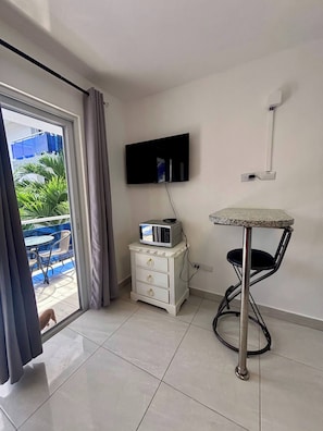 A room with a TV mounted on the wall, a small table with a chair, a white dresser, and a sliding glass door leading to an outdoor area with chairs and a blue fence. - Condo Neuf Avec Balcon Près de Tout à Sosua 206 (Sosúa)