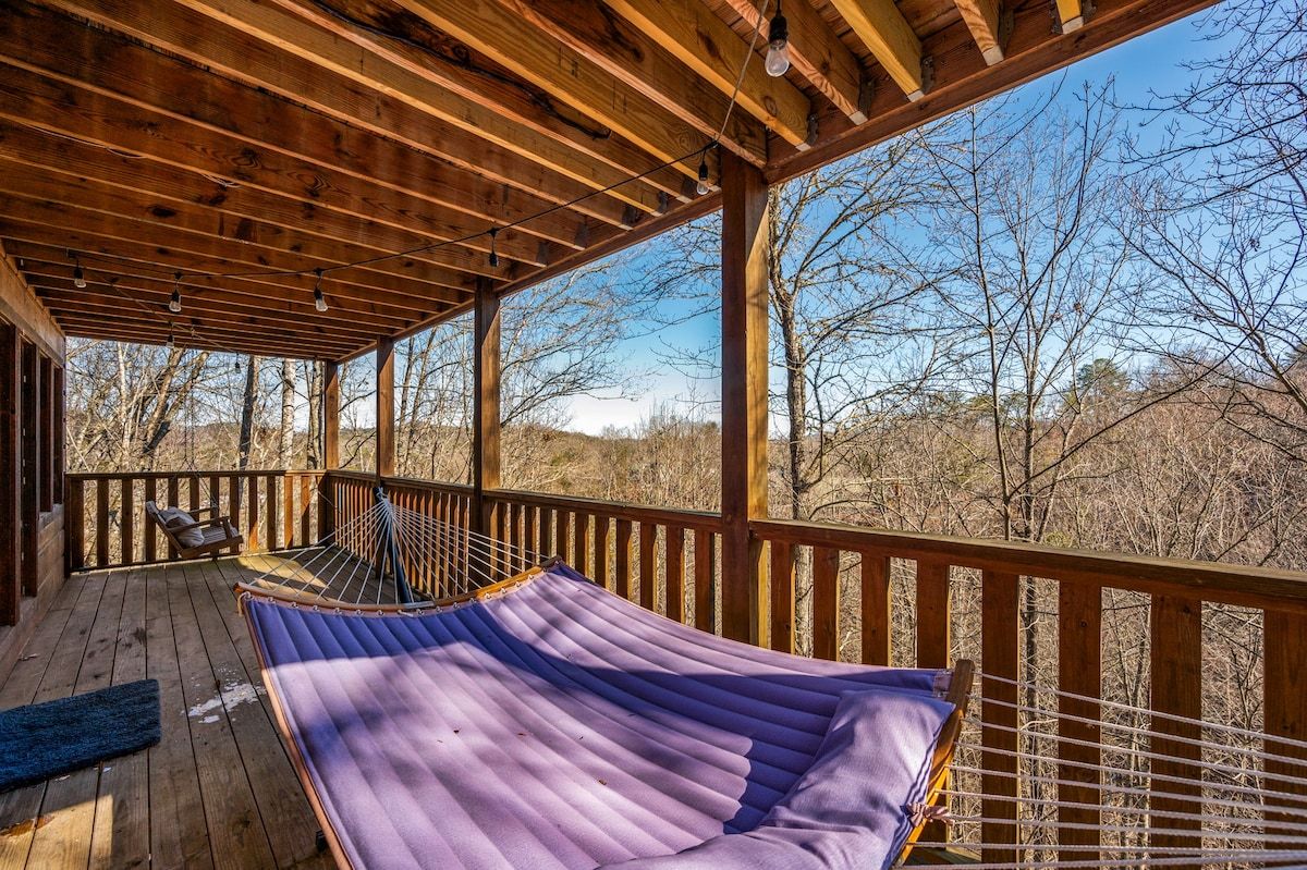 A wooden deck with a hammock and a hammock chair, overlooking a forested landscape.