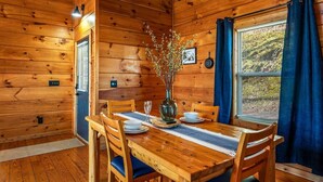 Un intérieur en bois avec une table à manger dressée pour deux personnes, un vase orné de branches et une fenêtre avec des rideaux.