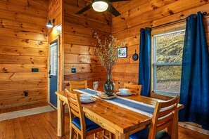 Un intérieur en bois avec une table à manger dressée pour deux personnes, un vase orné de branches et une fenêtre avec des rideaux.