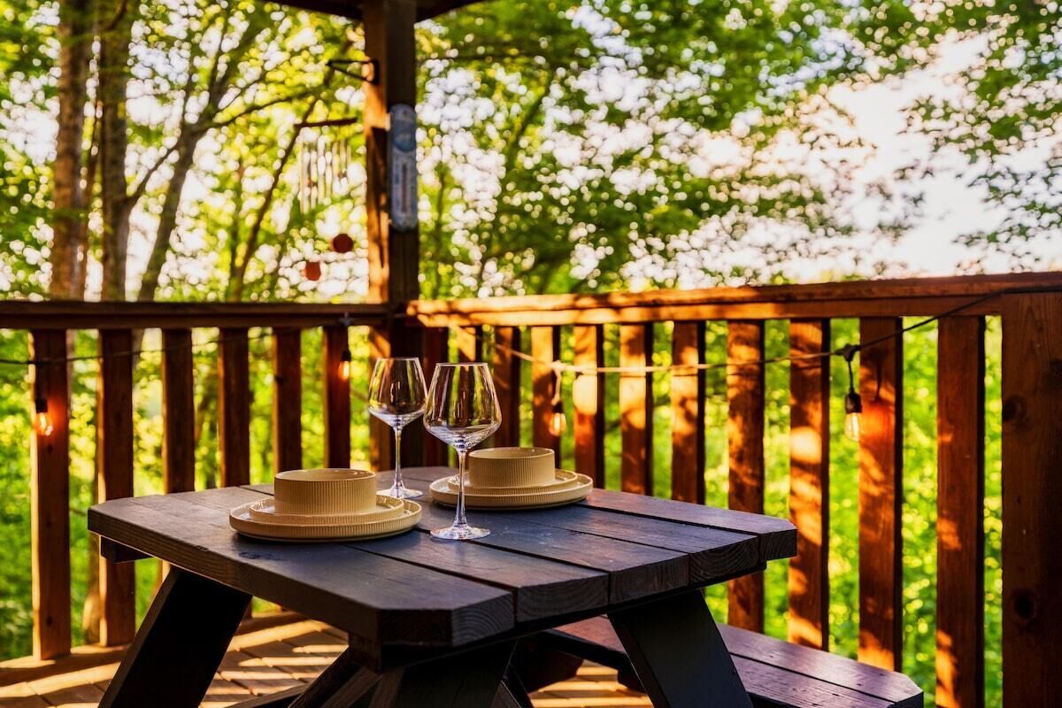 A wooden picnic table with two wine glasses and a hat on it, set against a backdrop of trees.