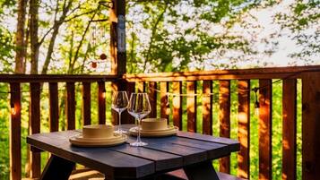 A wooden picnic table with two wine glasses and a hat on it, set against a backdrop of trees.