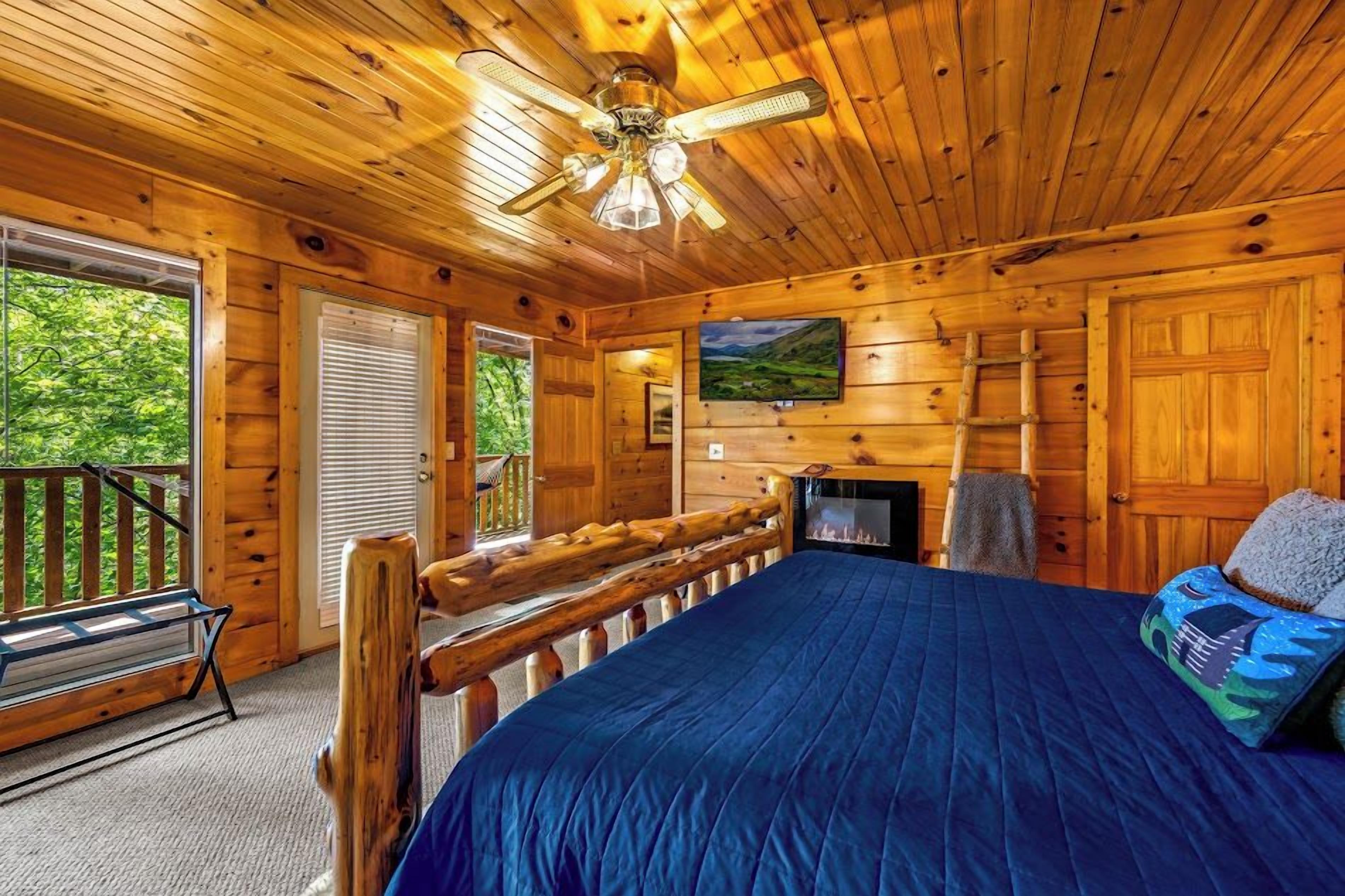 A log cabin bedroom with a bed, a ceiling fan, a fireplace, and a view of the outdoors.