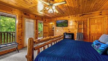 A log cabin bedroom with a bed, a ceiling fan, a fireplace, and a view of the outdoors.