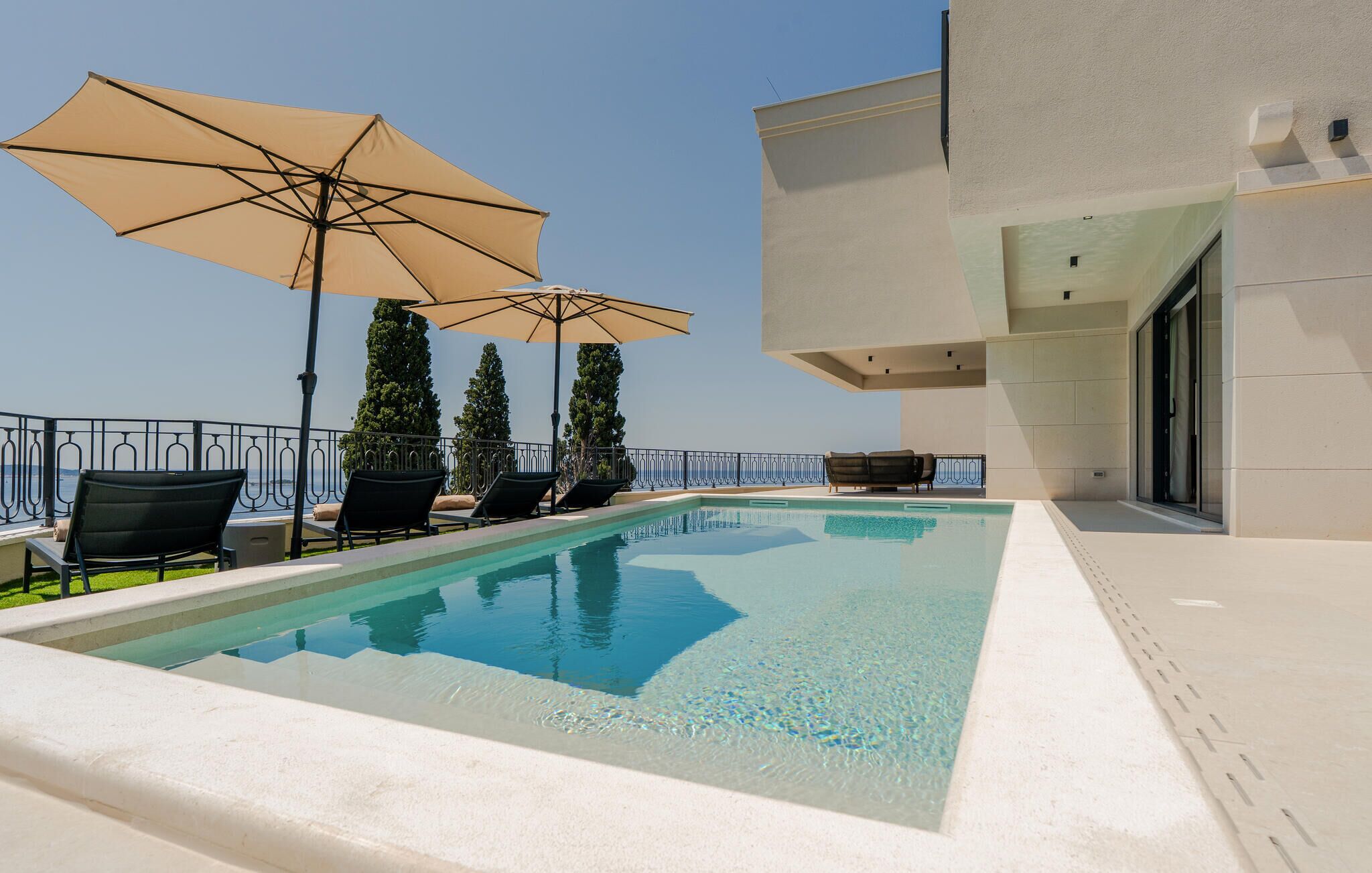 A swimming pool with lounge chairs and umbrellas, surrounded by a low fence and a building with large windows.