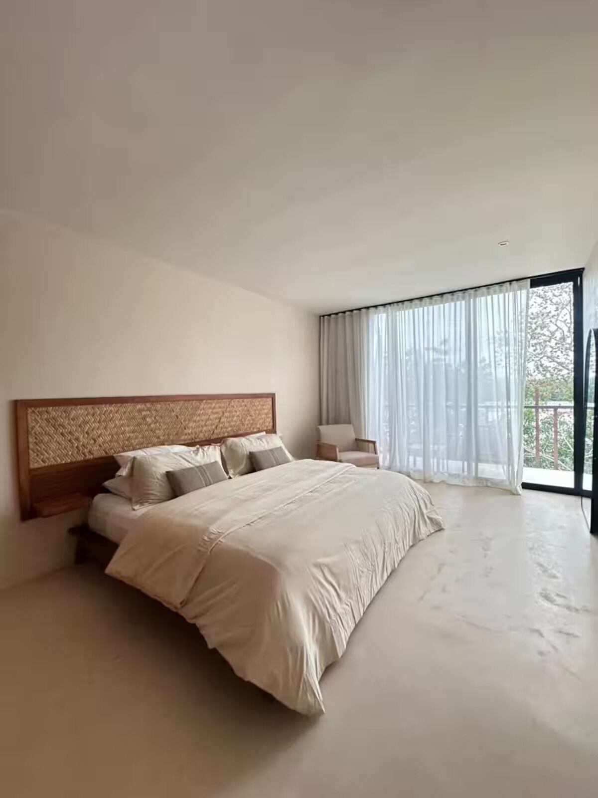 A modern bedroom with a large bed, a wooden headboard, a nightstand, and a view of greenery through sheer curtains.