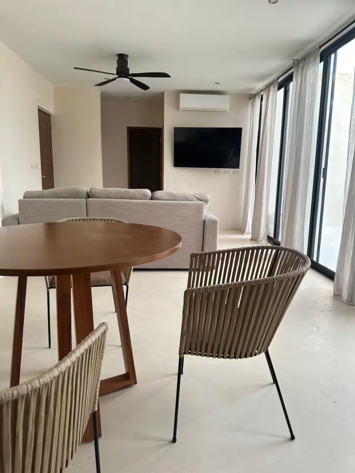 A modern living room with a round wooden table, two wicker chairs, a grey sofa, and a ceiling fan.