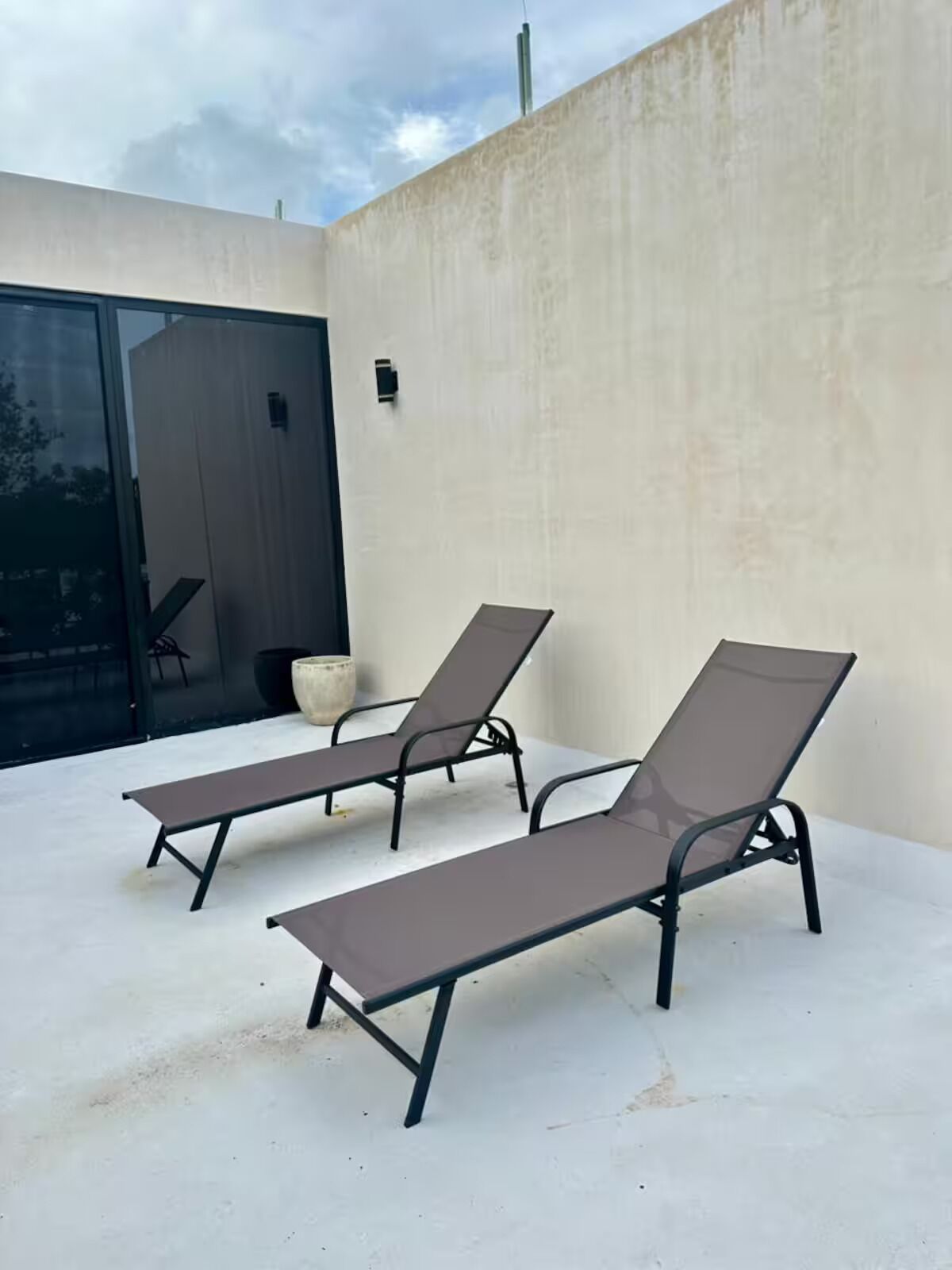 Two sun loungers with metal frames and dark cushions, positioned on a concrete patio adjacent to a building with a large glass door and a concrete wall.
