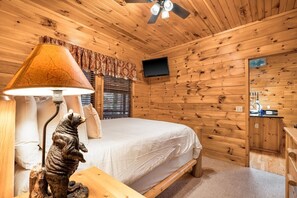 Et værelse i en træhytte med en seng, en lampe, en bjørnestatue, et fjernsyn og et vindue med persienner.