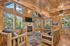 A log cabin interior with a stone fireplace, wooden furniture, and large windows. - Free Dollywood, Hot Tub, Firepit, Pool Table, Spa (Gatlinburg)
