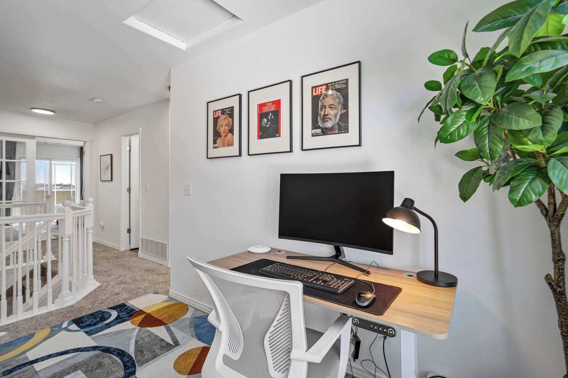 A modern office space with a desk, computer, and chair. There are three framed pictures on the wall, a potted plant, and a window with a view of the outside.