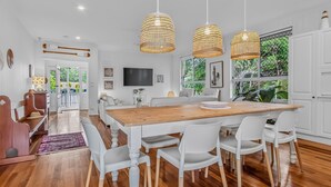 Ett rymligt matområde med ett träbord och vita stolar, omgivet av stora fönster som ger utsikt över grönskan.