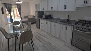 Cozinha moderna com armários brancos, fogão a gás, geladeira e área de jantar com mesa e cadeiras de vidro.
