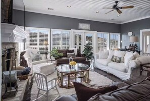 A living room with a fireplace, large windows, and a ceiling fan. - Lakefront Dream/Sleeps 16+/Pool/HotTub/Slides/Dock (Marble Falls)