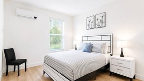 Une chambre à coucher comprenant un lit, des tables de chevet, une chaise, une fenêtre avec des stores et des œuvres d’art murales.