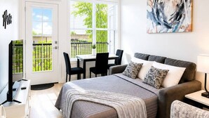 Une chambre à coucher moderne avec un lit, un canapé, un bureau et une chaise. Il y a une fenêtre avec des stores, une porte et un tableau au mur.