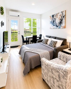 Een moderne slaapkamer met een bed, een bank, een bureau en een stoel. Er is een raam met zonwering, een deur en een schilderij aan de muur.