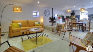 A modern living room with a sofa, coffee table, and chairs. There is a dining area with a table and chairs. The room has a large window and a clock on the wall.