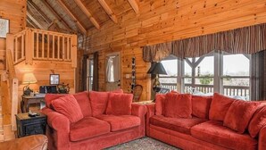 A cozy cabin interior with a red sofa, wooden walls, a window with a view, and a lamp on a table. - Barkwells Asheville Libby Cabin: Dog-Centric Resort, Hot Tub, Pond, Fenced Yard! (Mills River)