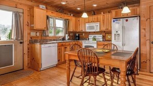 A wooden cabin with a kitchen featuring a dining table, chairs, a refrigerator, and a microwave. - Barkwells Asheville Libby Cabin: Dog-Centric Resort, Hot Tub, Pond, Fenced Yard! (Mills River)