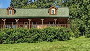 A log cabin with a green roof, a porch with railings, and dense surrounding greenery. - Barkwells Asheville Libby Cabin: Dog-Centric Resort, Hot Tub, Pond, Fenced Yard! (Mills River)