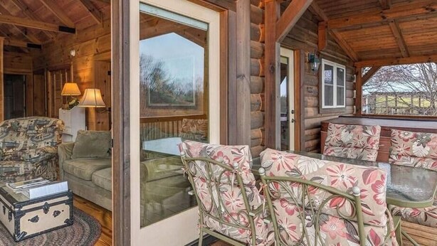 Una cabaña de madera con un porche, un sofá verde, sillas con estampado floral y una mesa de metal.