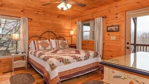 Un dormitorio de cabaña de madera con una cama, una mesita de noche, una ventana con cortinas, un ventilador de techo y vistas al exterior.