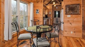 Interior de una cabaña de madera con una mesa redonda de comedor y cuatro sillas, una zona de cocina y un ventanal grande.