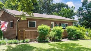 Une maison en bois d’un seul étage, avec un toit en pente, un porche à l’avant et une cour clôturée où poussent différentes plantes.