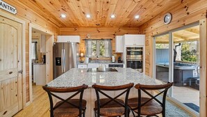 Interior de una cabaña de madera con cocina que incluye encimeras de granito, una isla central con taburetes y un amplio ventanal corredero que da a un área con hidromasaje al aire libre.