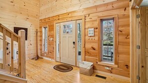 Interior de una cabaña de madera con una escalera, una puerta con ventana y un ventanal con vistas al exterior.
