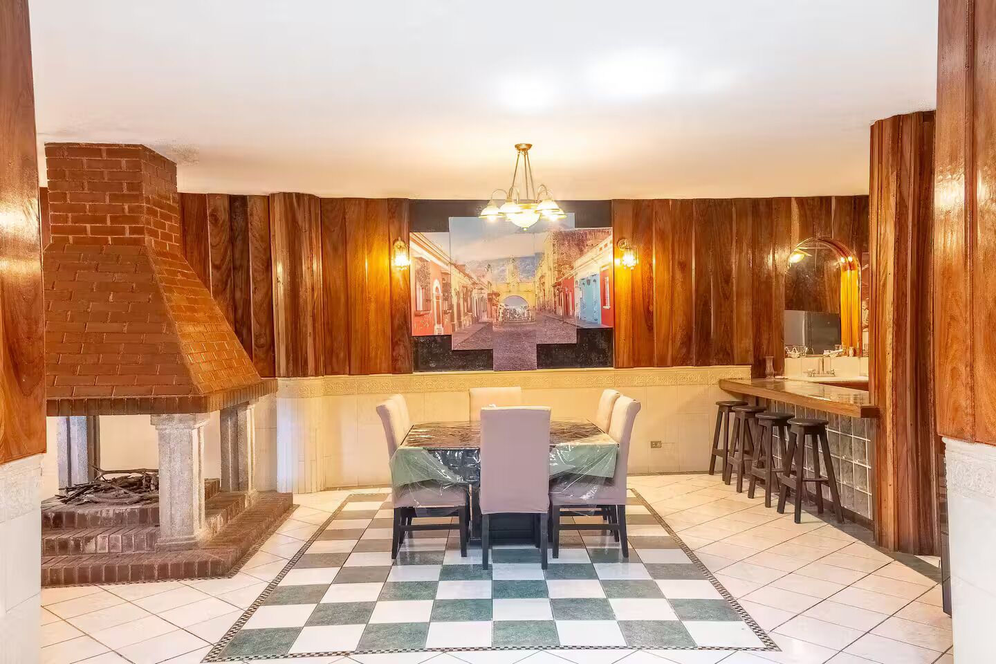 A dining area with a brick fireplace, wooden paneling, a checkered floor, and a bar with stools.