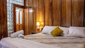 Een kamer met houten lambrisering, een bed met witte lakens en een geel kussen, een nachtkastje met een lamp, en een raam met geëtste glasschalen.