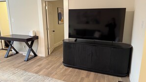 A living room with a flat-screen TV on a black stand, a dark wooden table, and a beige rug.