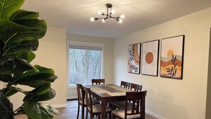 A dining area with a wooden table and chairs, a large window with a view of trees, and two framed artworks on the wall.