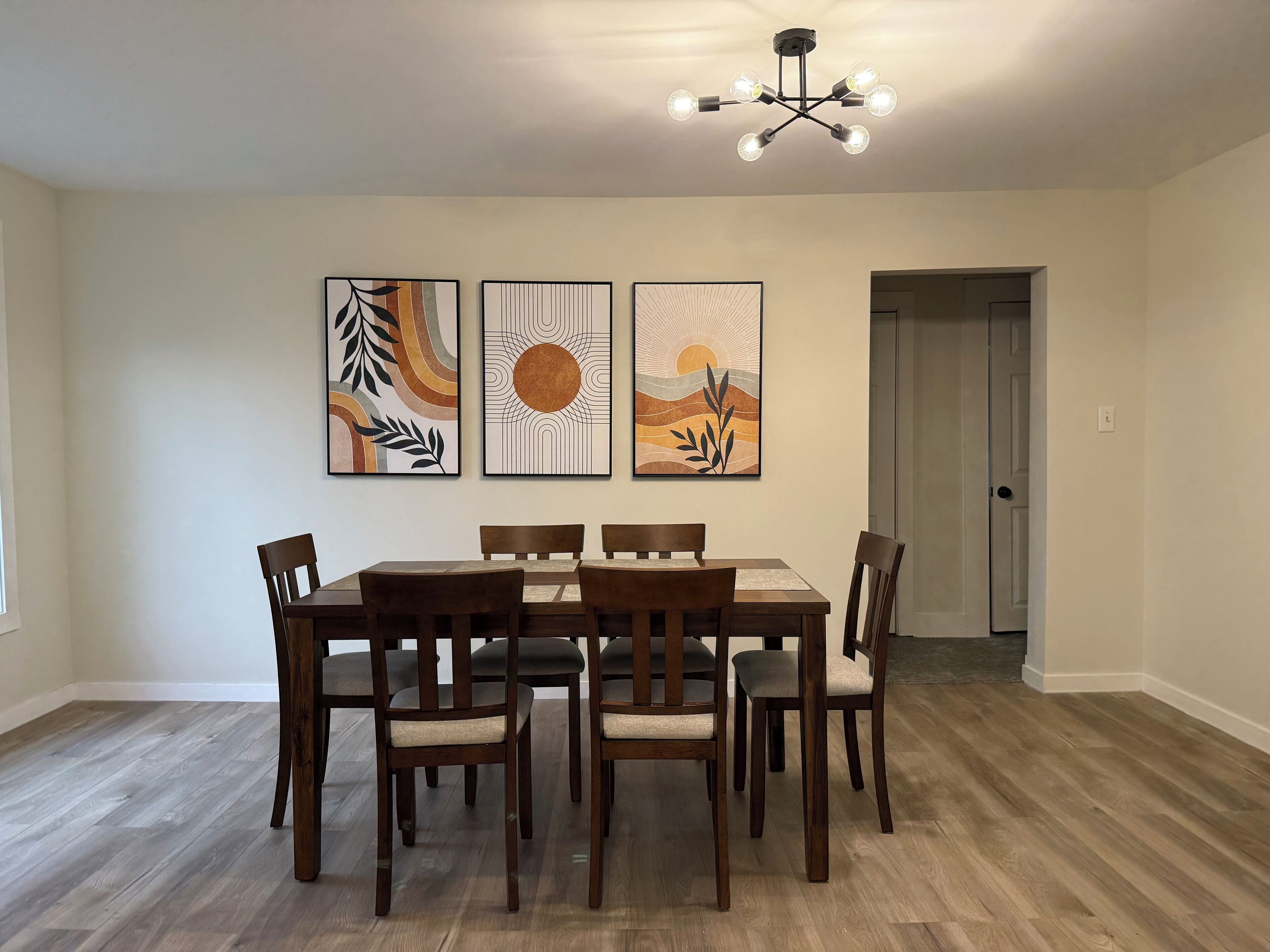 Un comedor con mesa y sillas de madera, tres obras de arte enmarcadas en la pared y una lámpara de techo moderna.