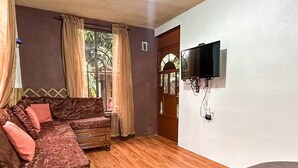 A cozy living room with a brown sofa, wooden flooring, a TV mounted on the wall, and a window with curtains. - Casa Familiar ubicada a minutos de Xetulul (Guatemala)
