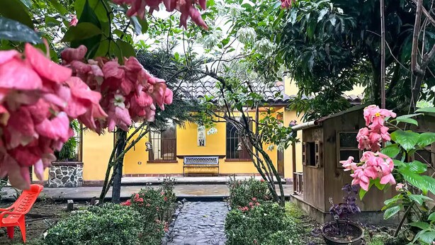 A garden pathway leading to a yellow building with a stone floor, surrounded by greenery and pink flowers. - Casa Familiar ubicada a minutos de Xetulul (Guatemala)