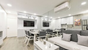 Een moderne woonkamer met een grijze bank, een witte salontafel, en een eethoek met witte tafels en stoelen.