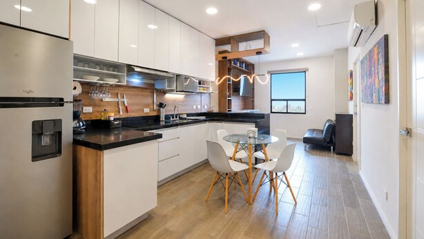 Een moderne keuken met een ingebouwde koelkast, een eethoek met een glazen tafel en witte stoelen, en een stadszicht door een groot raam.