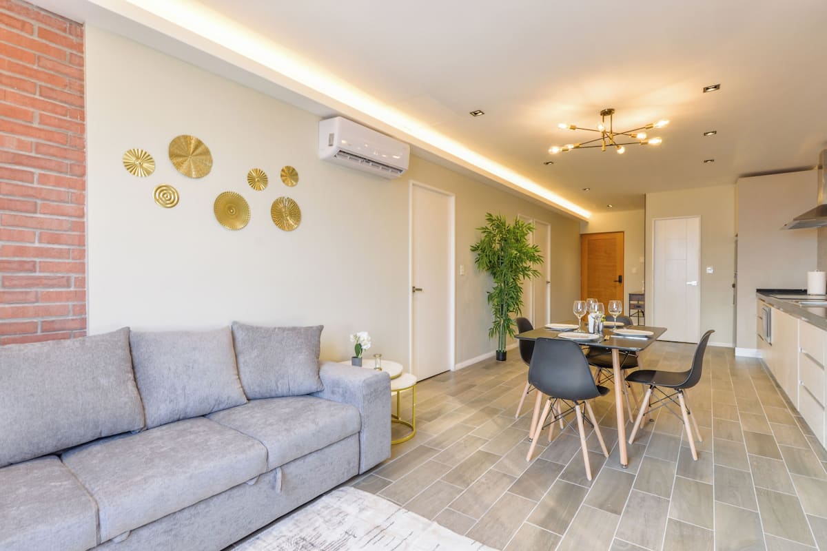A modern living room with a grey sofa, a dining area with a table and chairs, and a kitchen area with cabinets and a countertop.