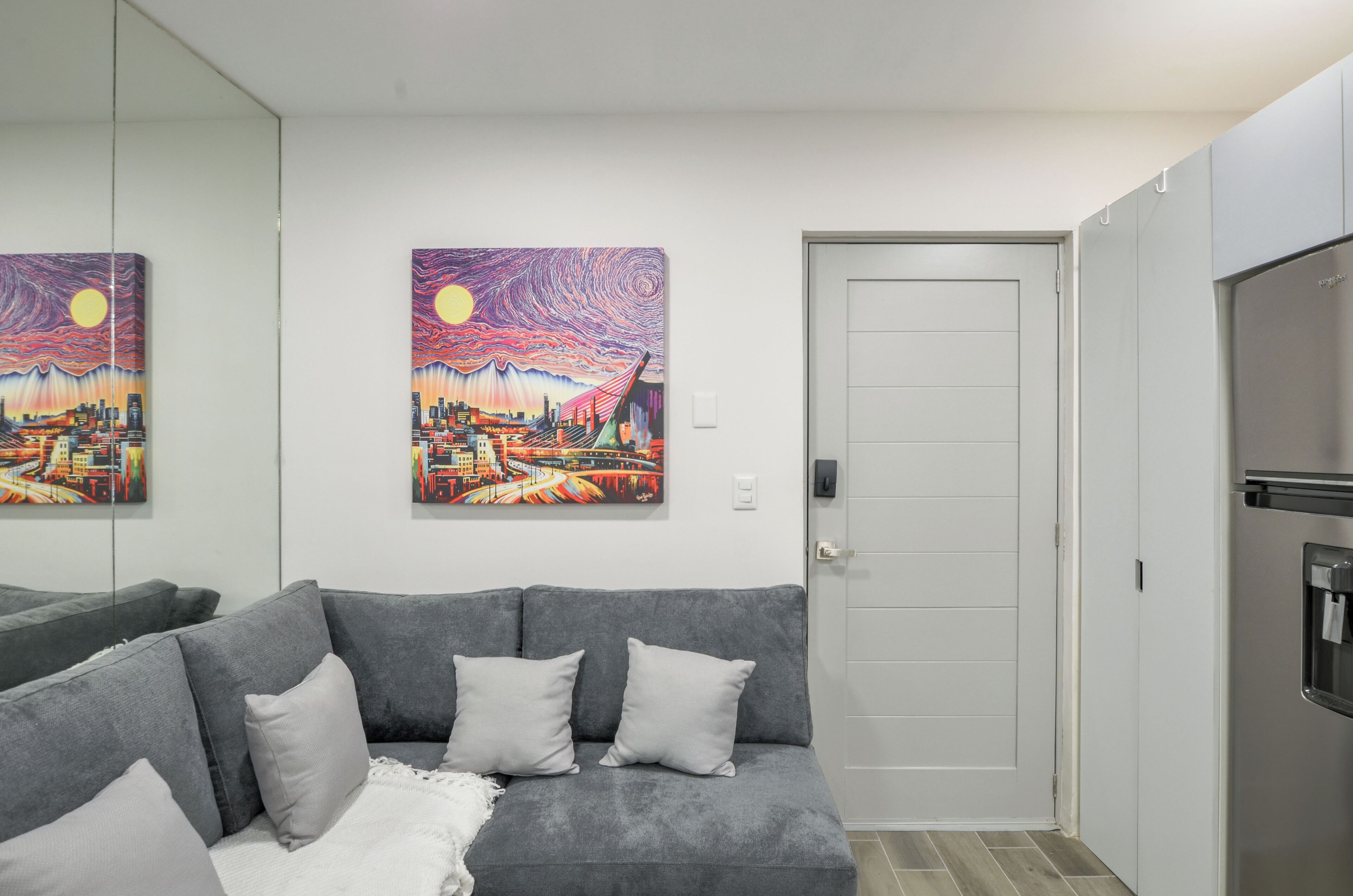 A living room with a grey sofa, two paintings, a white door, and a stainless steel refrigerator.