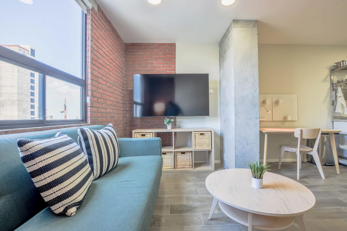 A modern living room with a blue sofa, a flat-screen TV mounted on a brick wall, a dining area with a table and chairs, and a large window offering a city view.