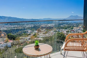 Un balcón con una mesa de vidrio y sillas de mimbre, con vistas a un paisaje urbano y montañas al fondo.