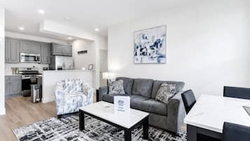 A modern living room with a grey sofa, a patterned armchair, a coffee table, and a dining table with chairs. There is a kitchen area with grey cabinets and a microwave. A painting hangs on the wall.