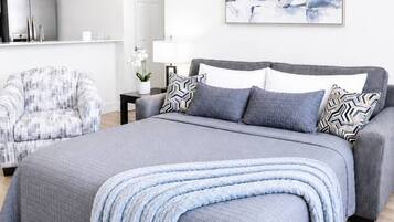 A modern bedroom with a large bed, a grey sofa, a patterned armchair, and a wall-mounted abstract painting.