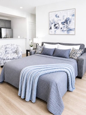 Een moderne slaapkamer met een groot bed, een grijze bank, een fauteuil met een patroon, en een abstract schilderij aan de muur.