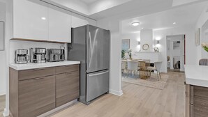 Cocina moderna con refrigerador de acero inoxidable, horno empotrado y microondas. Hay un mueble de madera con cajones y una encimera blanca. La cocina da a una zona de comedor con mesa y sillas blancas.